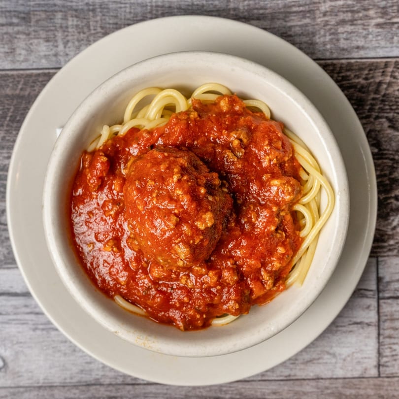 A white bowl of spaghetti with a large meatball and rich red marinara sauce.