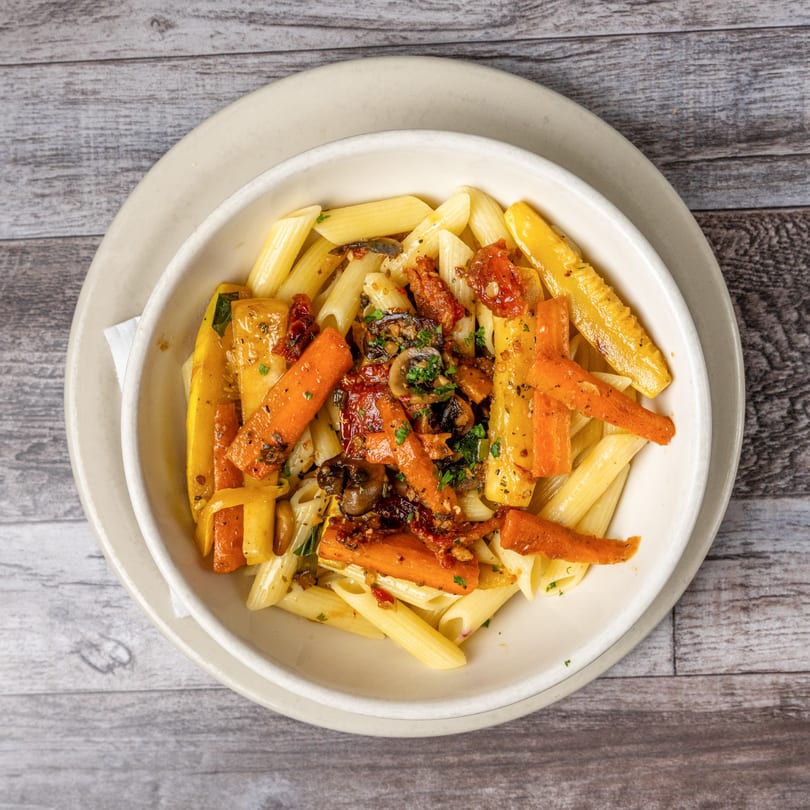 Penne pasta with roasted carrots, yellow squash, sun-dried tomatoes, and mushrooms in a white bowl.