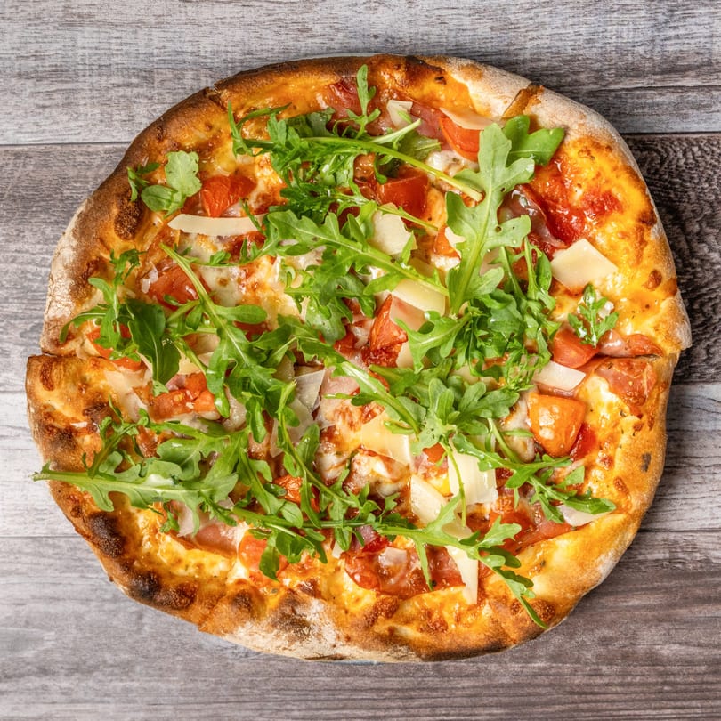 Round pizza with tomato sauce, mozzarella, prosciutto, arugula, and Parmesan cheese on a wooden surface.