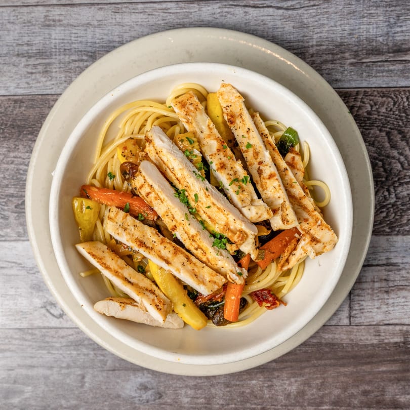 Overhead view of grilled chicken strips and roasted vegetables over spaghetti in a white bowl.
